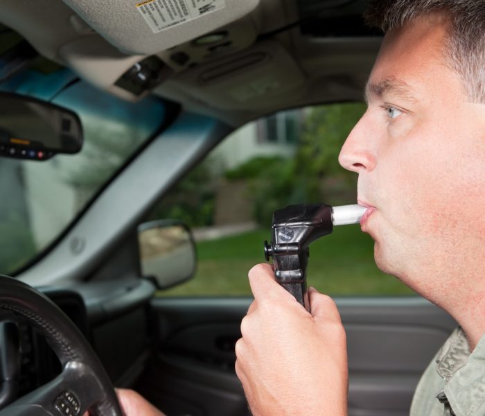 A male driver is blowing into an ignition interlock system which checks his alcohol concentration before allowing the vehicle to be started. A camera, mounted near the rear-view mirror, takes a picture of the driver to verify it is him blowing into the mouthpiece. Ignition interlock devices may be an alternative sentence for drunk driving or a probation requirement for those who have received a DUI.